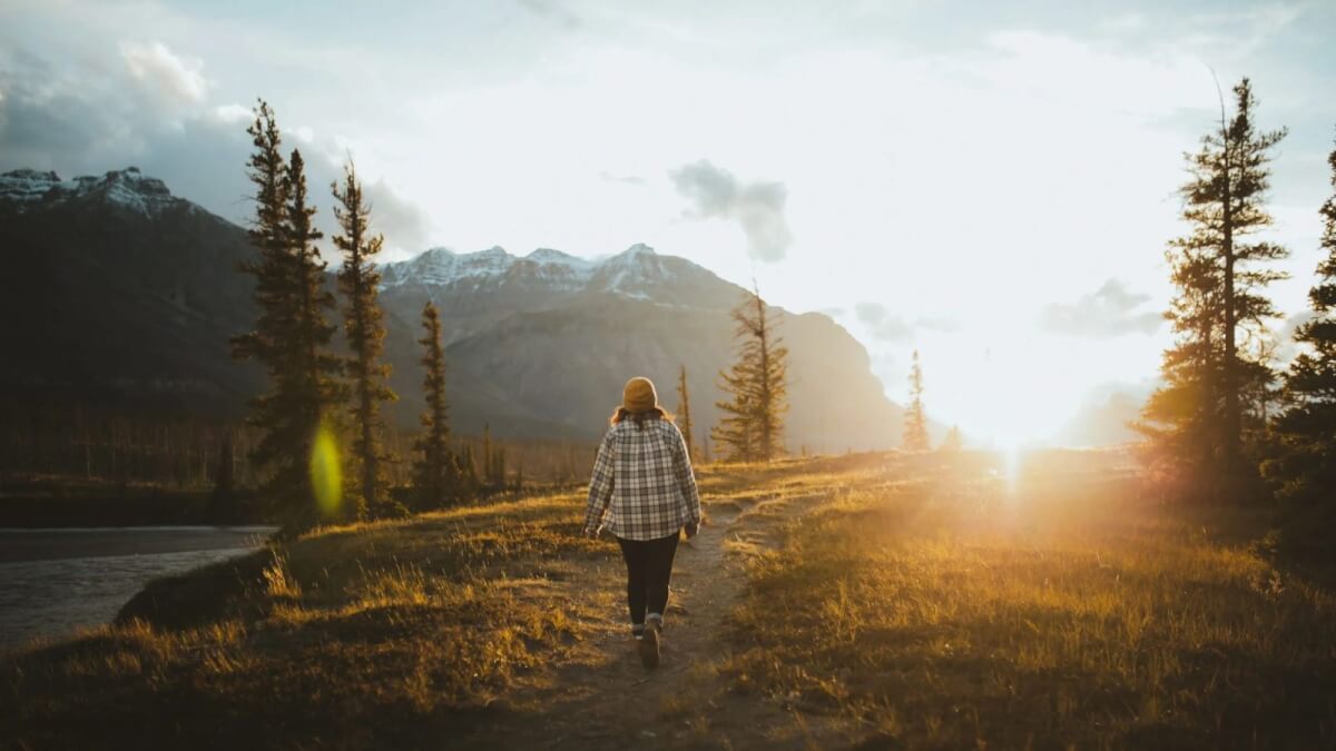 A person walks along a mountain trail toward the setting sun, surrounded by pine trees and wilderness, illustrating how time in nature can support the grieving process.