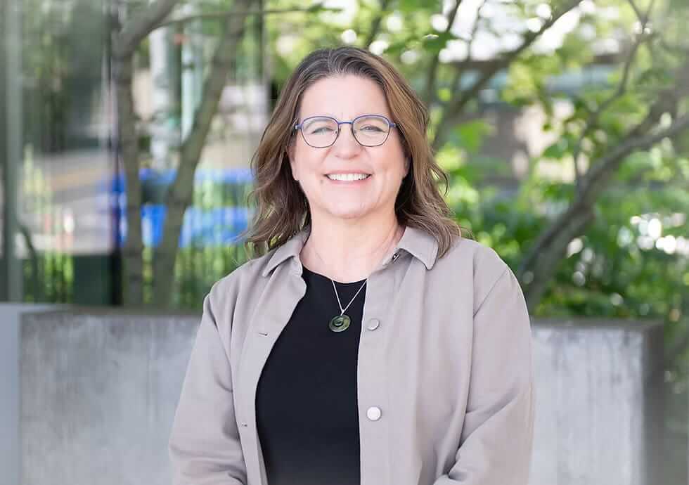Author headshot of Sarah Cavanaugh, writer and creator of the book and podcast Peaceful Exit, smiling outdoors with glasses, a grey button-up shirt, and a black undershirt against a blurred green backdrop.
