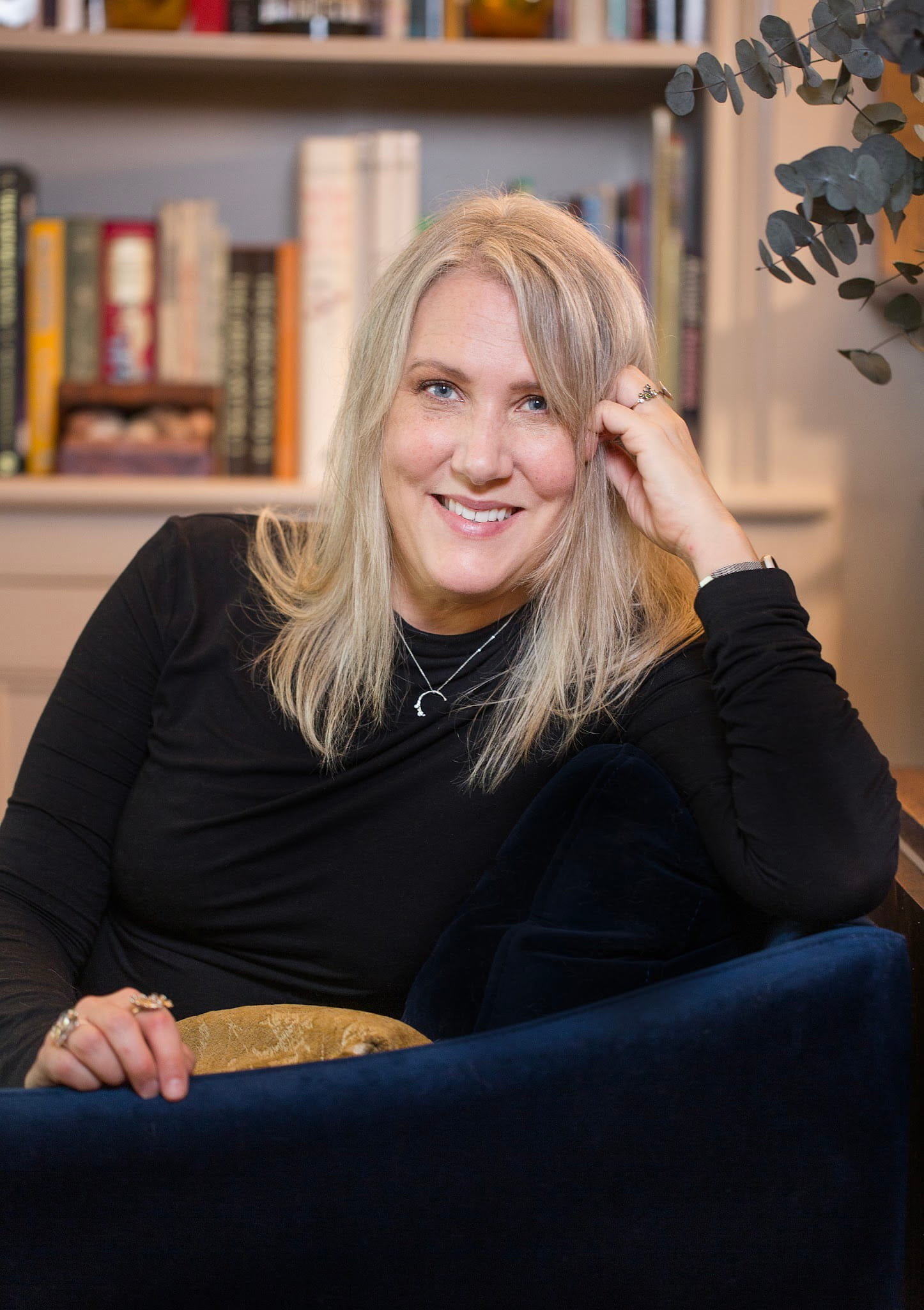 Smiling blonde woman in a black long-sleeve shirt sitting on a blue velvet chair in front of a bookshelf.