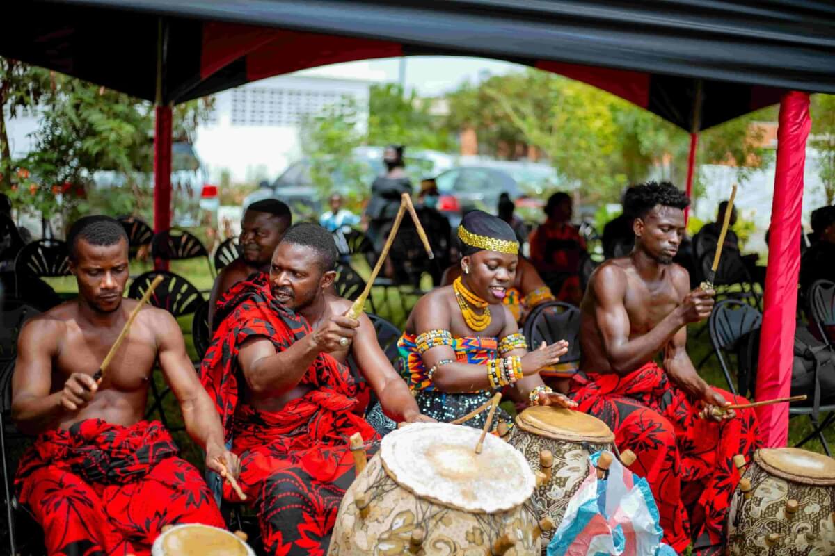 Musicians in traditional red and black attire play drums under a canopy at a Ghanaian celebration of life funeral.