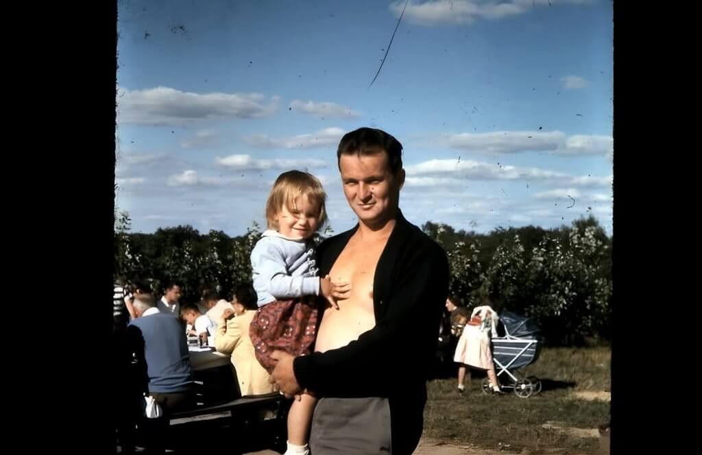 Color photograph of a young Richard holding one of his four daughters outdoors at a family gathering, capturing a moment from their life together.